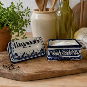 May include: A white ceramic butter dish with a lid featuring blue hand-painted floral patterns and the text "Huamantla" in black script. The rectangular base is filled with white butter, and the lid is placed beside it on a wooden surface.
