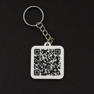 Op de afbeelding: Een witte vierkante sleutelhanger met een zwart QR-code ontwerp. De sleutelhanger is bevestigd aan een zilveren metalen ring en ketting. De achtergrond is een donker, getextureerd oppervlak.