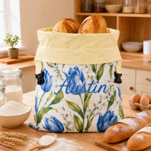 Peut inclure: Un sac à pain en tissu avec un motif de tulipes bleues et de feuilles vertes sur fond blanc. Le prénom Austin est brodé en bleu sur le devant. Le sac possède un revers supérieur jaune pâle et deux boucles latérales en plastique noir.