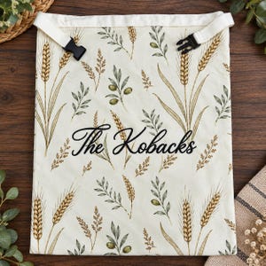 May include: A cream-colored fabric bag featuring a repeating pattern of golden wheat stalks and green olive branches. The bag has a white top border with black plastic side-release buckles and the text The Kobacks in black script.