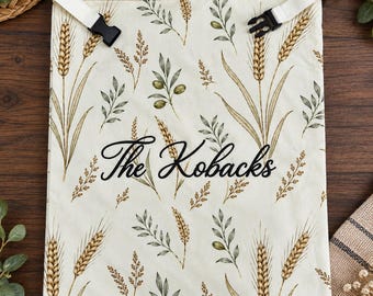 May include: A cream-coloured fabric bag featuring a repeating pattern of golden wheat stalks and green olive branches. The bag has a white top border with black plastic side-release buckles and the text The Kobacks in black script.