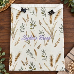 May include: A cream-colored fabric bag featuring a repeating pattern of wheat stalks and olive branches. The bag has white straps with black plastic side-release buckles at the top. The text Sophia's Bread is embroidered in purple.