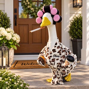 May include: A white goose figurine wearing a leopard print bathrobe with the name Olivia embroidered on the chest. The goose has pink and black hair rollers on its head, a cigarette in its beak, and holds a small orange slice and a coffee cup.
