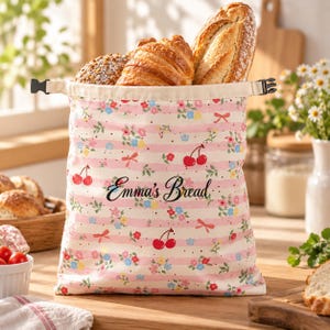 May include: A fabric bread bag featuring a pink and white striped pattern with small floral motifs and red cherry illustrations. The bag has a black plastic buckle closure at the top and the text "Emma's Bread" printed in black script on the front.