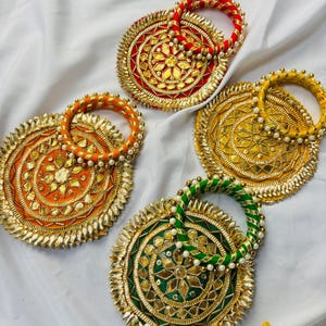Könnte beinhalten: Drei runde Stoffuntersetzer und passende Armreifen in Rot, Orange, Gelb und Grün. Jeder Untersetzer verfügt über ein zentrales sternförmiges Spiegeldesign, goldene Stickereien und einen Rand aus kleinen, spitzen, goldfarbenen Akzenten.