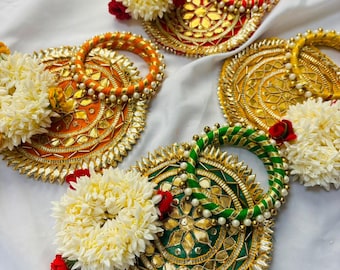 May include: Four circular decorative plates featuring intricate gold-toned metallic patterns and mirror accents. Each plate is paired with a matching fabric-wrapped bangle and adorned with a cluster of white jasmine-like flowers and small red roses.