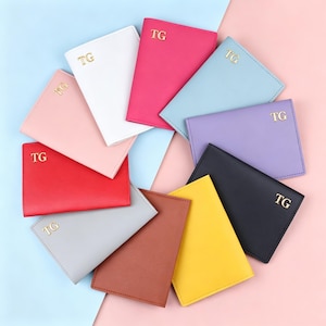 May include: A collection of colorful passport holders in various shades, including pink, red, white, blue, purple, black, brown, yellow, and gray. Each holder is embossed with the gold letters "TG". The holders are arranged on a pink and blue background.