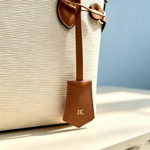May include: A white bucket bag with a textured, wavy surface pattern. The bag features brown leather accents, including a hanging luggage tag embossed with the gold-colored initials JK, and brown leather straps attached with gold-tone hardware.