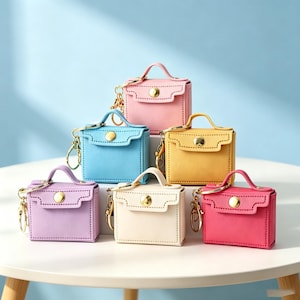 Könnte beinhalten: Eine Sammlung von sechs Miniatur-Handtaschen-Schlüsselanhängern in Rosa, Hellblau, Gelb, Lavendel, Creme und Magenta. Jede kleine Tasche verfügt über einen Tragegriff, einen Klappenverschluss mit goldenem Druckknopf und einen Metallschlüsselring.