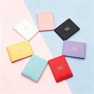 May include: A collection of personalized wallets in various colors, including white, pink, black, yellow, purple, teal, and red. Each wallet features the gold-colored initials "TG" on the front. The wallets are arranged in a circular pattern against a two-tone pink and blue background.