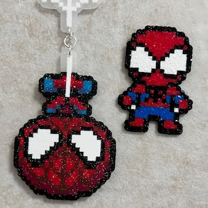 May include: A pixel art style keychain featuring a round red and blue character head with white eyes, attached to a white glittery web-shaped charm, alongside a separate small pixel art figure of a red and blue character with large white eyes.