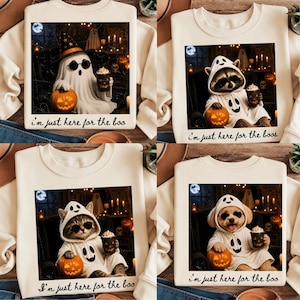 Op de afbeelding: Vier crèmekleurige sweatshirts met grafische prints van een geest, een wasbeer, een kat en een hond, elk in een geestkostuum, met een pompoen en een beker, voorzien van de tekst "i'm just here for the boo" of "i'm just here for the boos".