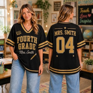 Op de afbeelding: Een zwart shirt met V-hals en gouden strepen op de mouwen en zoom. De voorkant bevat de tekst "FOURTH GRADE" en "Mrs. Smith" met een icoon van een boek en potlood. De achterkant toont "MRS. SMITH" en het nummer "04" met gouden sterren.