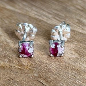May include: A pair of stud earrings featuring round, vibrant magenta and white mottled stones set in four-prong silver metal settings. Each earring includes a matching silver metal butterfly-style clutch back, providing a secure fit for the post.