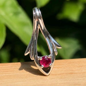 May include: A silver pendant featuring a teardrop-shaped frame with a polished metallic finish. A single, round, deep pinkish-red gemstone is securely held in a four-prong setting at the base of the open, elongated, and pointed metal structure.