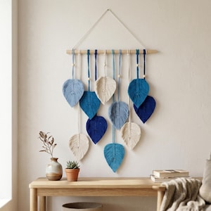 May include: A macrame wall hanging featuring ten leaf-shaped fiber art pieces in shades of cream, light blue, and dark blue. The leaves are suspended from a horizontal wooden dowel by cotton cords, accented with small wooden beads throughout.
