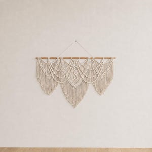 May include: A cream-colored macrame wall hanging suspended from a horizontal wooden dowel. The piece features intricate knotted patterns, multiple draped swags, and long, loose fringe that forms three distinct pointed sections along the bottom edge.