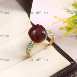 Puede incluir: Un anillo de oro con una gran piedra preciosa de color rojo oscuro tallada en forma de cojín facetado en el centro. La banda está decorada con pequeñas piedras de color azul brillante incrustadas en los laterales. AuraMilan es visible.