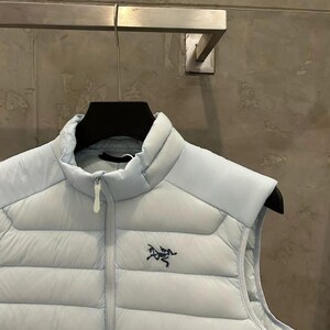 May include: A light blue puffer vest with a high collar and a zipper closure. The vest features a small, dark blue logo on the chest. The vest is hanging on a black hanger.