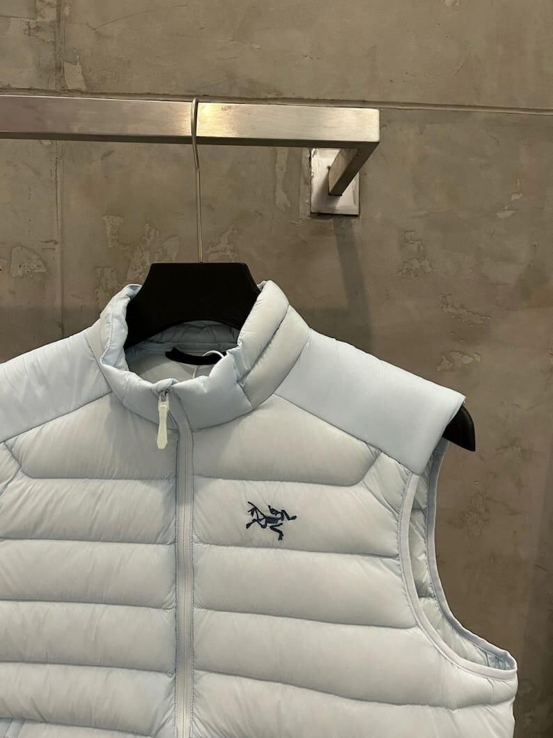 May include: A light blue puffer vest with a high collar and a zipper closure. The vest features a small, dark blue logo on the chest. The vest is hanging on a black hanger.