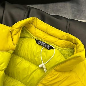 May include: Bright yellow puffer jacket with a quilted design. The jacket has a high collar and a tag with the brand name "Arc'teryx". The jacket is made of a smooth, shiny material. The background is a dark, textured surface.