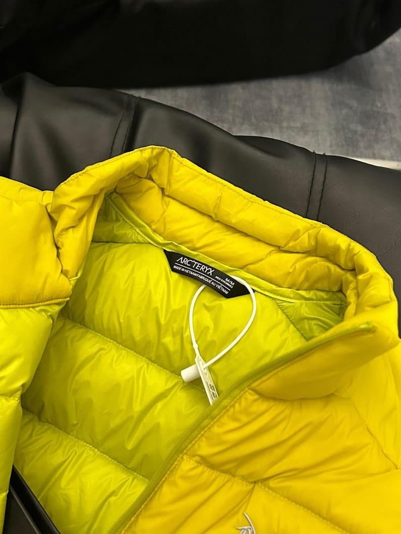 May include: Bright yellow puffer jacket with a quilted design. The jacket has a high collar and a tag with the brand name "Arc'teryx". The jacket is made of a smooth, shiny material. The background is a dark, textured surface.