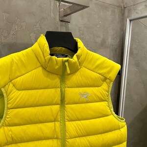 May include: A bright yellow puffer vest with a high collar and a front zipper. The vest has a quilted design and a small logo on the chest. The vest is sleeveless and appears to be made of a lightweight material.