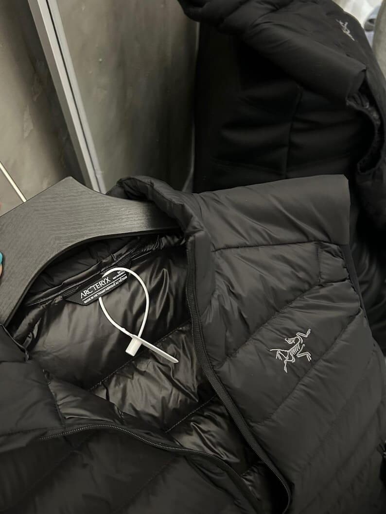 May include: Black quilted down jacket with a shiny interior. The jacket features a zipper closure and a small logo on the chest. The jacket is hanging on a black hanger.