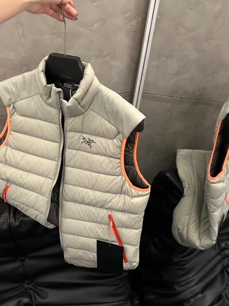 May include: A light gray puffer vest with orange trim and zippers. The vest is sleeveless and has a high collar. The Arc'teryx logo is visible on the chest. The vest is hanging on a black hanger.