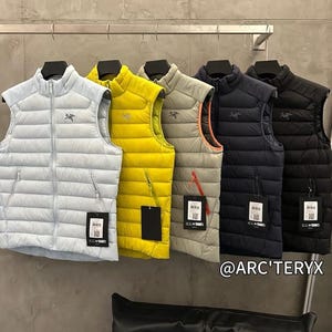 May include: A row of five puffer vests in various colors: light blue, yellow, gray, navy, and black. Each vest has a high collar, a zipper, and a small logo. The vests are hanging on a metal rack.