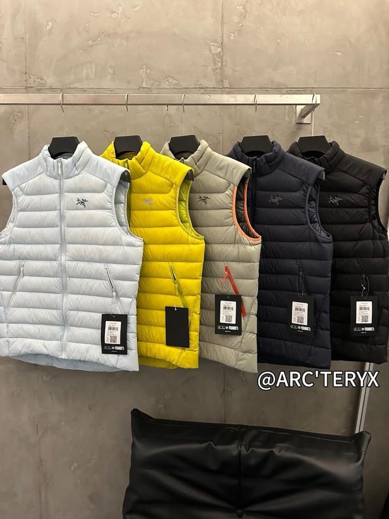 May include: A row of five puffer vests in various colors: light blue, yellow, gray, navy, and black. Each vest has a high collar, a zipper, and a small logo. The vests are hanging on a metal rack.