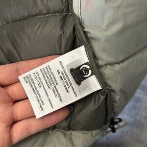 May include: Close-up of a gray and olive green vest with a tag. The tag includes text in multiple languages, including "CERIUM VEST M" and "100% NYLON". The vest has a quilted design and a drawstring.