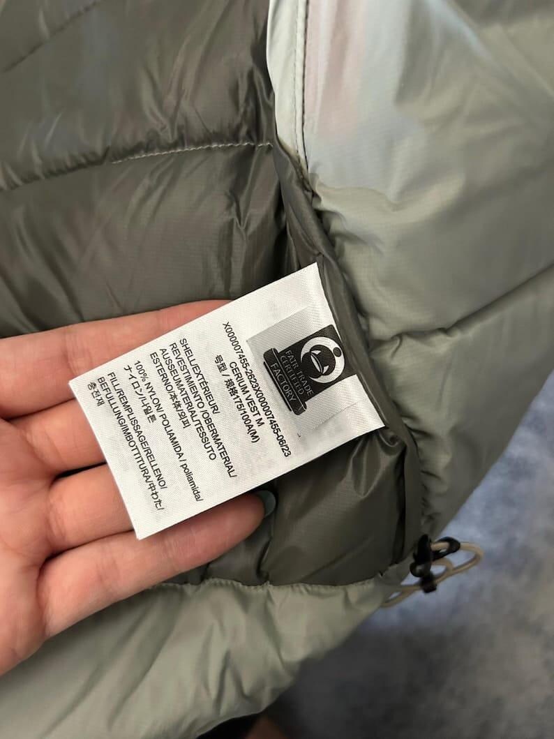 May include: Close-up of a gray and olive green vest with a tag. The tag includes text in multiple languages, including "CERIUM VEST M" and "100% NYLON". The vest has a quilted design and a drawstring.