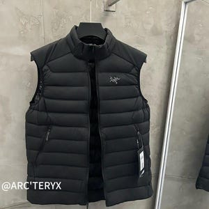 May include: Black puffer vest with a high collar and a full-length zipper. The vest has two side pockets and a small logo on the chest. The brand name is visible at the bottom left corner. The vest is hanging on a black hanger.