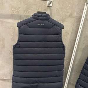 May include: Navy blue puffer vest with a quilted design. The vest has a stand-up collar and the brand name "ARCTERYX" printed on the back. The vest is hanging on a metal hanger against a neutral background.