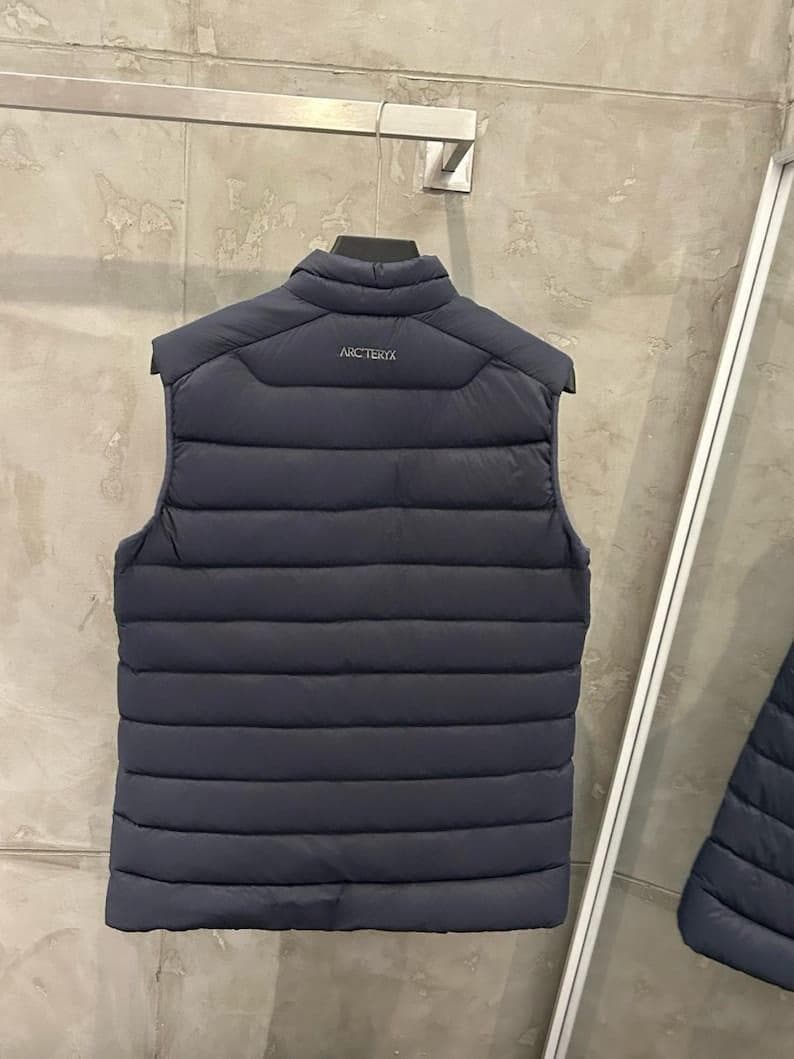 May include: Navy blue puffer vest with a quilted design. The vest has a stand-up collar and the brand name "ARCTERYX" printed on the back. The vest is hanging on a metal hanger against a neutral background.