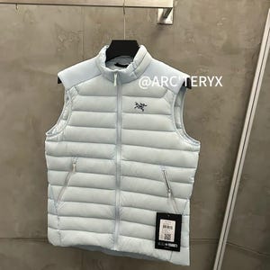 May include: A light blue puffer vest with a high collar and a zipper closure. The vest has two side pockets and a small logo on the chest. The text "@ARCTERYX" is visible. A tag with a barcode hangs from the vest.