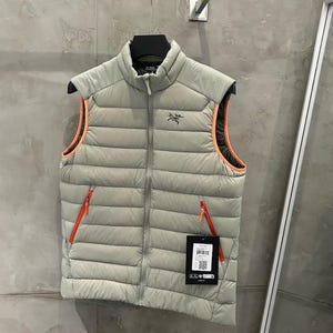 May include: A light gray puffer vest with orange trim and zippers. The vest has a high collar and a small logo on the chest. It is hanging on a black hanger against a gray wall.