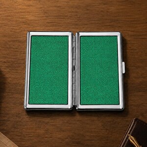 May include: Open, silver-framed business card holder with a textured green interior. The card case is displayed open on a wooden surface, with a pen and notebook in the background.