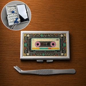 Op de afbeelding: Een rechthoekige metalen pillendoos met een vintage cassettebandje-afbeelding en bloemrijke randen, vergezeld van een zilveren pincet. De doos is gesloten en in een inzetstuk geopend afgebeeld, met witte en blauwe pillen erin.