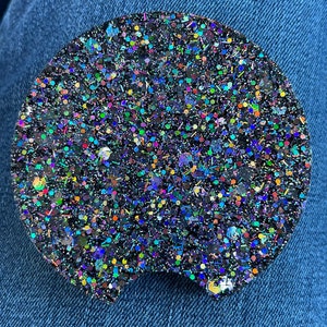 May include: A black round coaster with iridescent glitter embedded in the surface. The coaster has a slightly irregular shape with a rounded bottom edge.