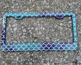 Pode incluir: Um porta-placas de carro de glitter azul-esverdeado e roxo com um design de escamas de sereia.