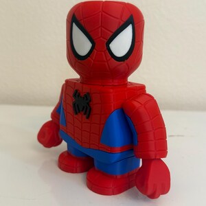 May include: A red and blue Spider-Man figurine. The figure has a red head with white eyes and a black outline. The body is red with a black spider emblem on the chest, blue arms and legs, and red hands and feet. The figurine is a collectible.