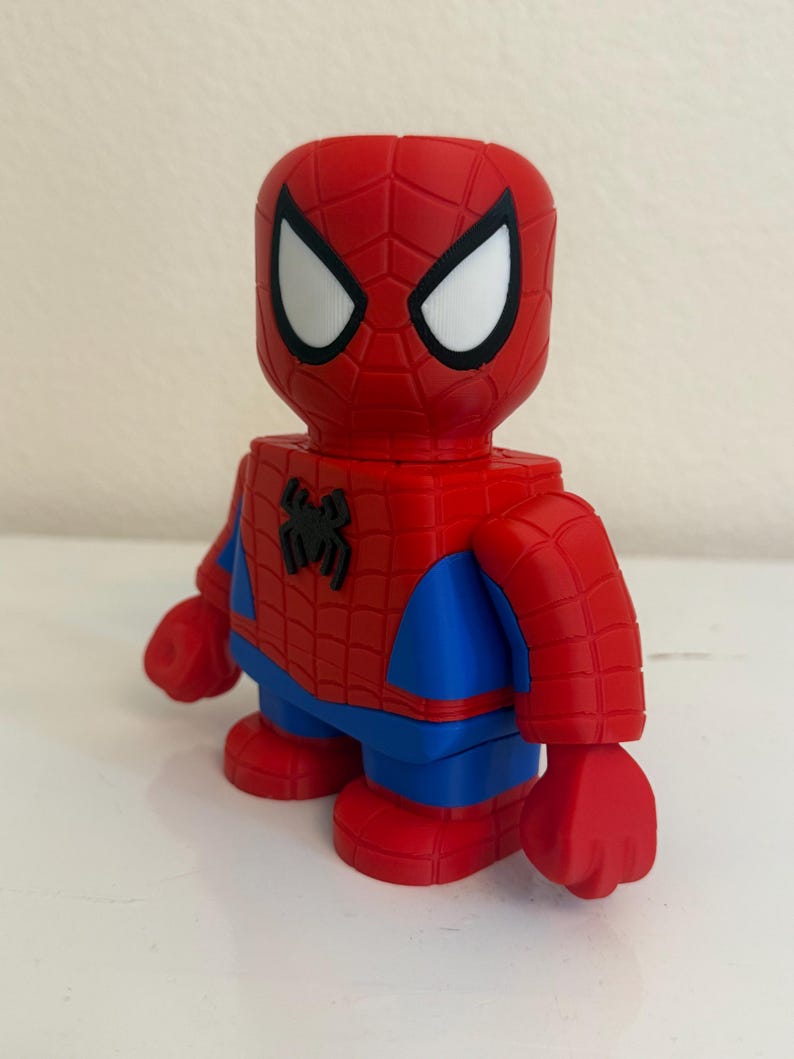May include: A red and blue Spider-Man figurine. The figure has a red head with white eyes and a black outline. The body is red with a black spider emblem on the chest, blue arms and legs, and red hands and feet. The figurine is a collectible.
