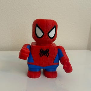 May include: A red and blue Spider-Man figurine with a blocky design. The figure has white eye shapes, a black spider emblem on the chest, and blue accents. The toy is standing upright.