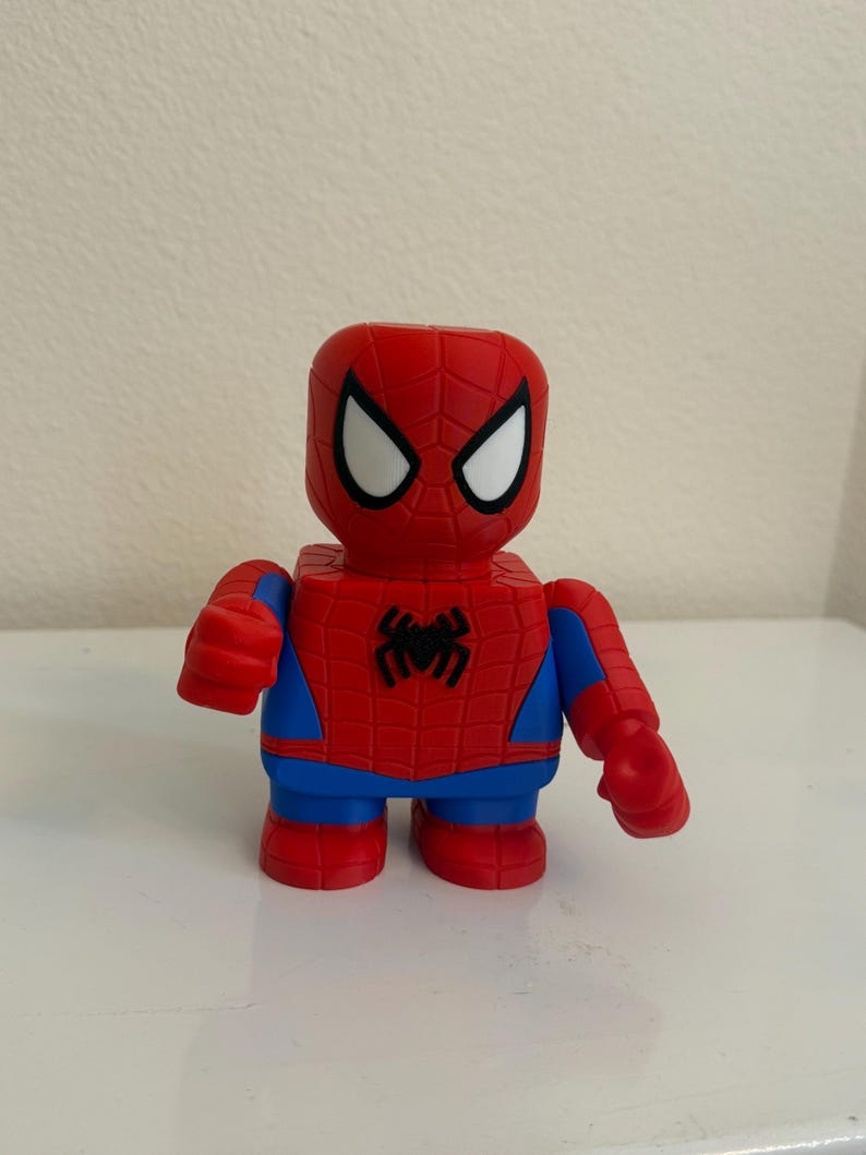 May include: A red and blue Spider-Man figurine with a blocky design. The figure has white eye shapes, a black spider emblem on the chest, and blue accents. The toy is standing upright.