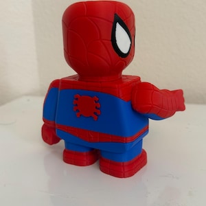 May include: A red, blue, and white Spider-Man figurine. The figure has a red head and arms, a blue torso and legs, and red feet. The Spider-Man figure is in a blocky, cartoon style.