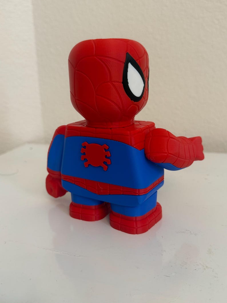 May include: A red, blue, and white Spider-Man figurine. The figure has a red head and arms, a blue torso and legs, and red feet. The Spider-Man figure is in a blocky, cartoon style.