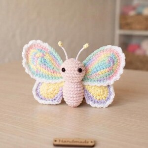 May include: A crocheted butterfly toy featuring a light pink body and head with two dark eyes and antennae. The wings are crafted in pastel rainbow colors including yellow, blue, and purple, finished with a white scalloped edge border.