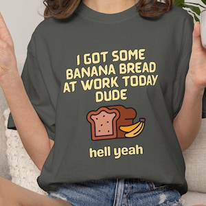 Puede incluir: Una camiseta de manga corta gris carbón con texto amarillo que dice I GOT SOME BANANA BREAD AT WORK TODAY DUDE y hell yeah, con una ilustración gráfica de una rebanada de pan de plátano marrón y dos plátanos amarillos en el centro.